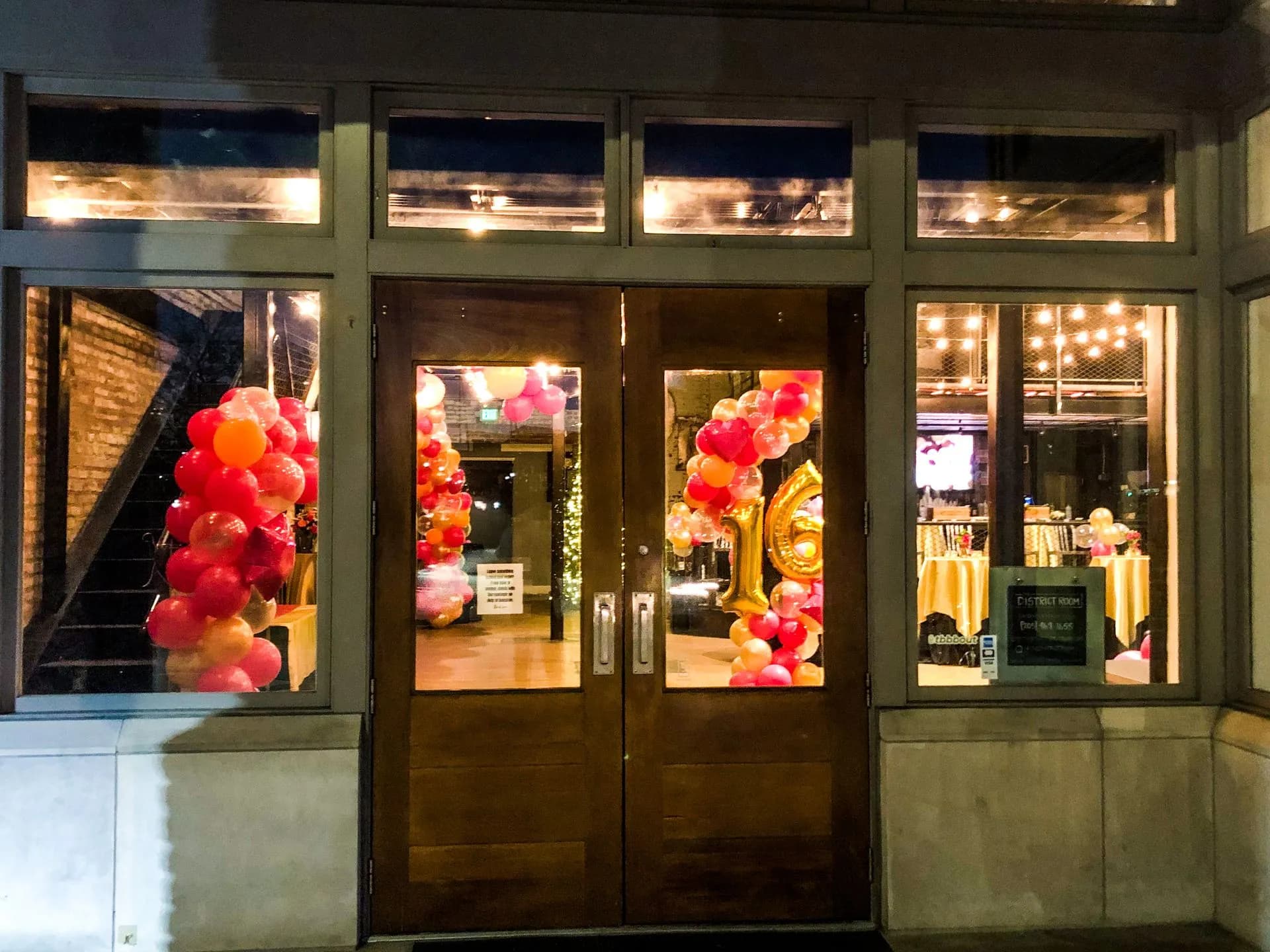 The District Room's front entrance at night, with balloon garlands and string lights glowing through the windows